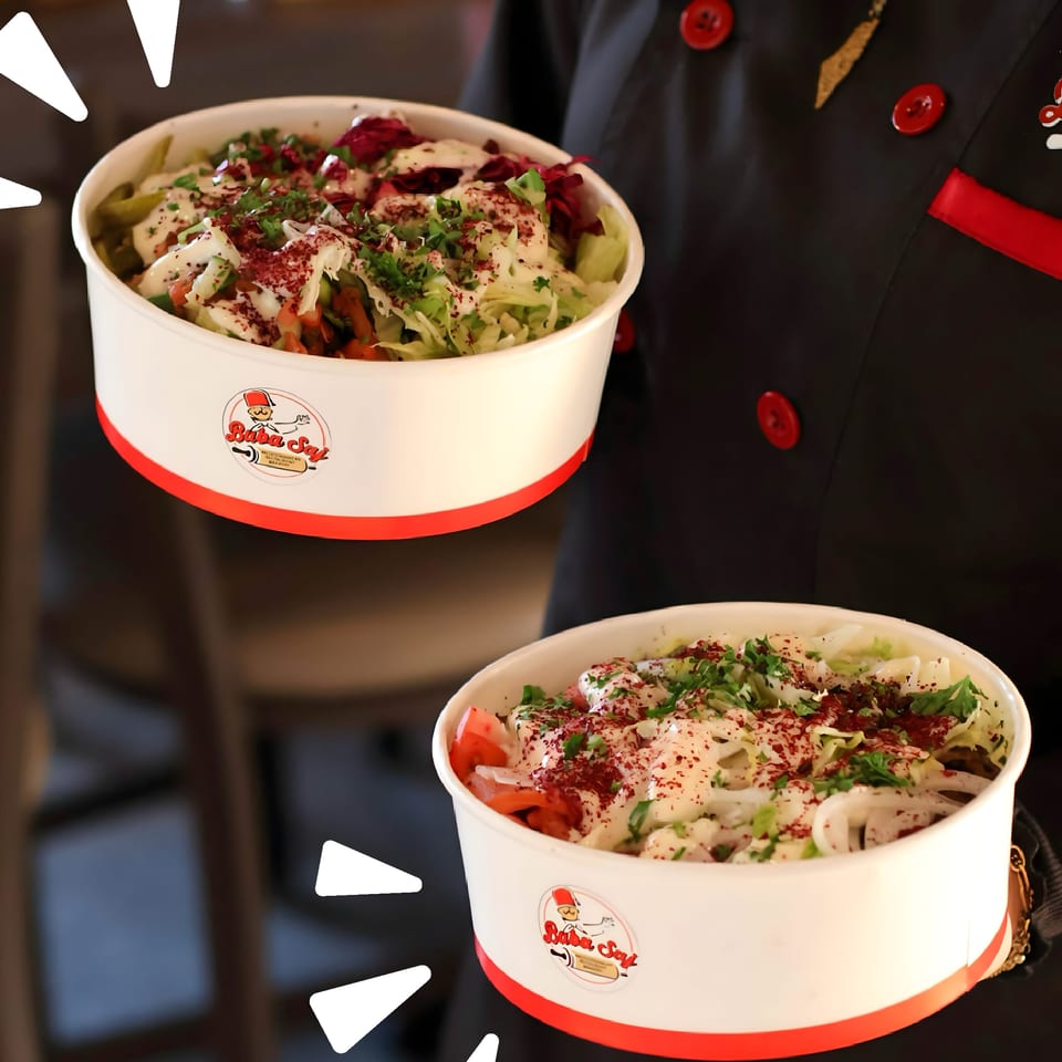 Two Buba Say bowls filled with shawarma, lettuce, tomatoes, herbs, creamy sauce, and red spices, held by a chef in a black uniform.