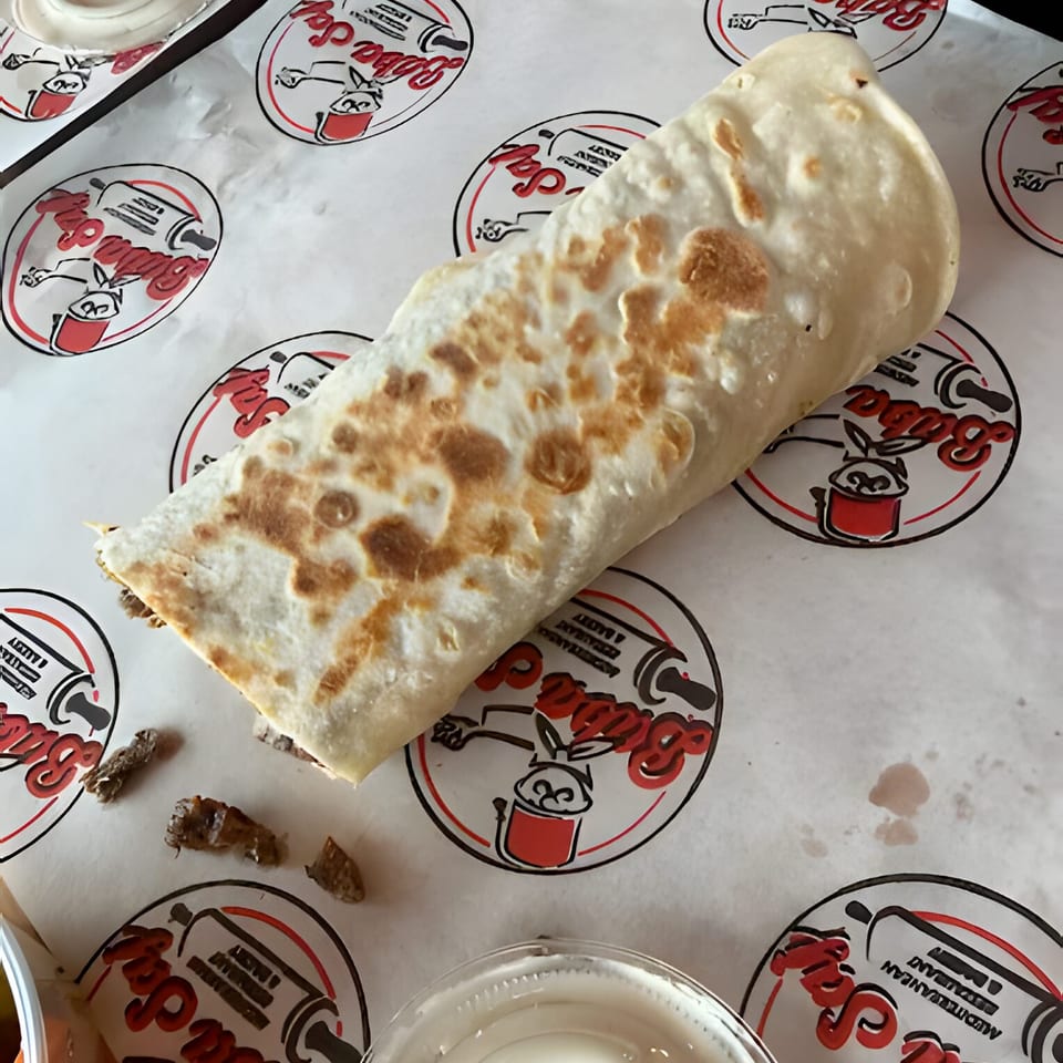 A browned, folded flatbread wrap rests on branded paper beside small dishes of creamy sauce.